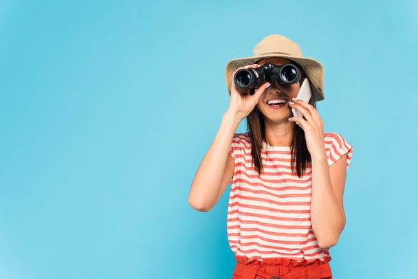 happy woman in hat talking on smartphone and looking through binoculars isolated on blue - Foto, Imagem