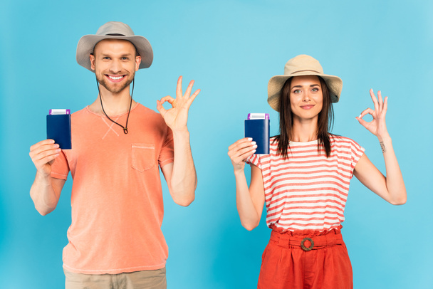szczęśliwy mężczyzna i kobieta w kapeluszach posiadających paszporty i pokazując ok znak na niebieski - Zdjęcie, obraz