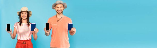 πανοραμική φωτογραφία του ευτυχισμένου ζευγαριού σε καπέλα κρατώντας smartphones με λευκή οθόνη και διαβατήρια που απομονώνονται σε μπλε  - Φωτογραφία, εικόνα