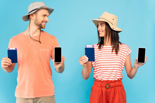 szczęśliwa para w kapeluszach trzymających smartfony z pustym ekranem i paszportami patrząc na siebie odizolowanych na niebiesko  - Zdjęcie, obraz
