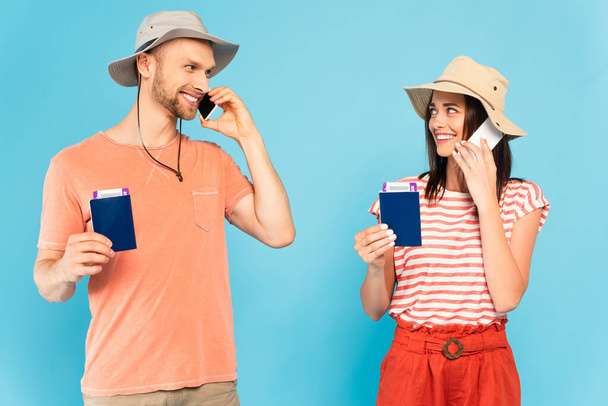 feliz pareja en sombreros hablando en teléfonos inteligentes, sosteniendo pasaportes y mirándose aislados en azul  - Foto, Imagen