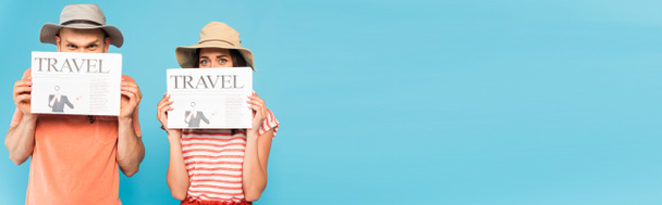 panoramic crop of couple in hats covering faces with travel newspapers isolated on blue  - Photo, Image