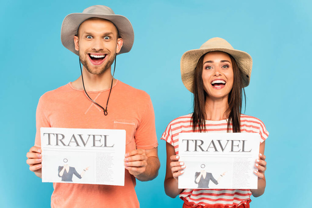 podekscytowana para w kapeluszach trzymających gazety podróżnicze i patrzących na kamerę odizolowaną na niebiesko  - Zdjęcie, obraz