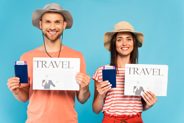 happy couple in hats holding travel newspapers and passports isolated on blue  - Foto, afbeelding