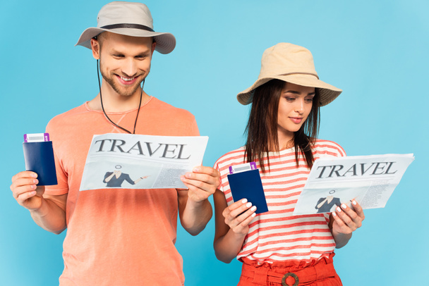 happy couple in hats reading travel newspapers and holding passports isolated on blue  - Foto, Imagem