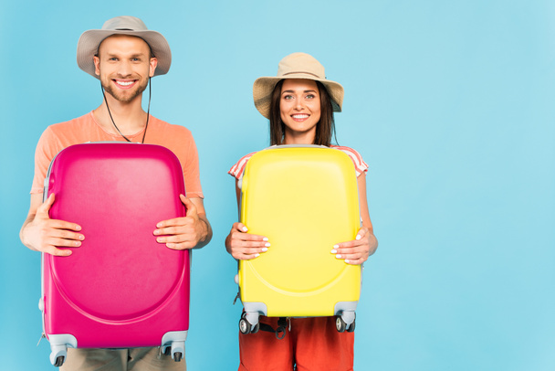 happy couple looking at camera and holding luggage isolated on blue  - Photo, Image