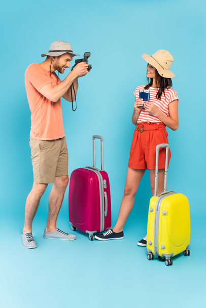 hombre feliz en el sombrero de tomar la foto de la chica con pasaportes mientras sostiene la cámara vintage y de pie cerca de equipaje en azul - Foto, Imagen