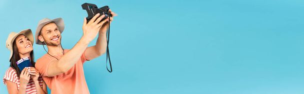 panoramiczny strzał szczęśliwy człowiek w kapeluszu gospodarstwa vintage aparat fotograficzny i selfie z wesołą dziewczyną odizolowany na niebiesko - Zdjęcie, obraz