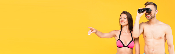 strona nagłówka wesołej kobiety wskazując palcem podczas gdy mężczyzna bez koszulki patrząc przez lornetkę odizolowany na żółtym  - Zdjęcie, obraz