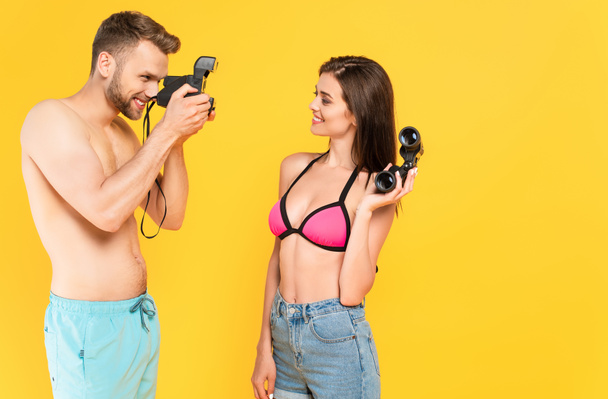 cheerful man taking photo of girl in swimsuit holding binoculars isolated on yellow  - Zdjęcie, obraz