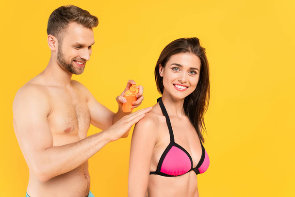 muscular man smiling while applying sunblock on cheerful woman isolated on yellow - Fotografie, Obrázek