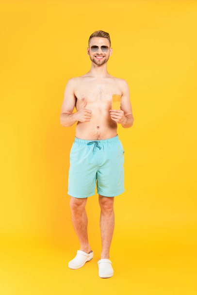 happy shirtless man in sunglasses showing thumb up and holding bottle with sunblock on yellow  - Foto, afbeelding