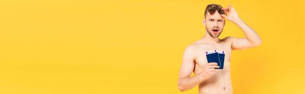 cultivo panorámico de hombre impactado y sin camisa tocando gafas de sol y sosteniendo pasaportes aislados en amarillo  - Foto, Imagen
