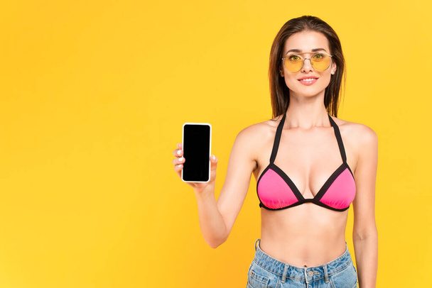 wesoła dziewczyna w okularach przeciwsłonecznych gospodarstwa smartfon z pustym ekranem izolowane na żółtym - Zdjęcie, obraz