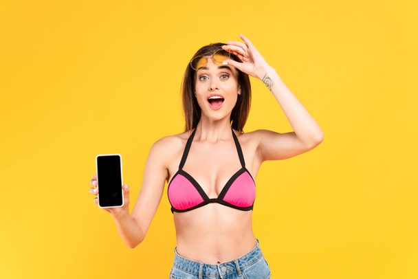 aufgeregtes Mädchen im Badeanzug berührt Sonnenbrille und hält Smartphone mit leerem Bildschirm isoliert auf gelb - Foto, Bild