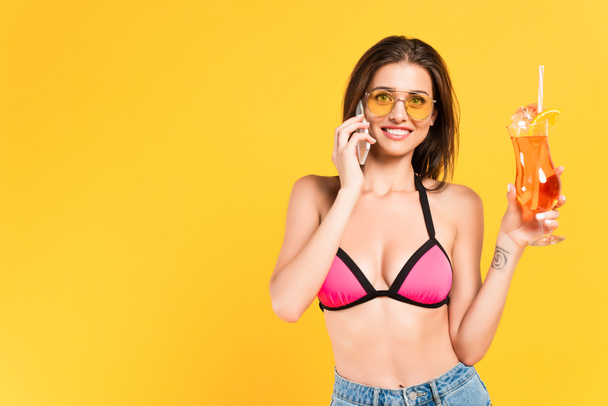 happy girl in sunglasses talking on smartphone and holding cocktail isolated on yellow - Photo, image