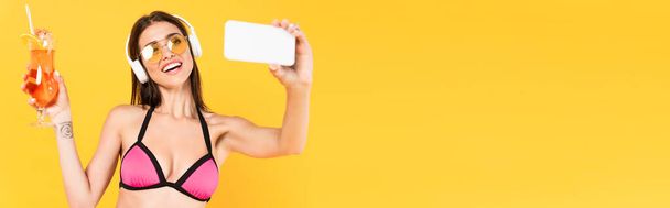 concept panoramique de fille joyeuse et tatouée dans des lunettes de soleil prendre selfie tout en tenant cocktail isolé sur jaune  - Photo, image