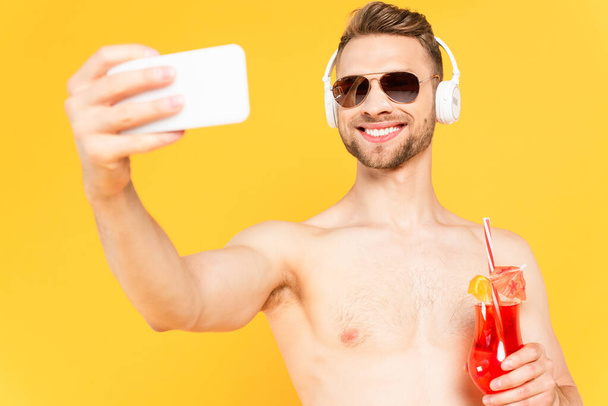 enfoque selectivo de hombre alegre y sin camisa en gafas de sol y auriculares tomando selfie mientras sostiene cóctel aislado en amarillo  - Foto, Imagen