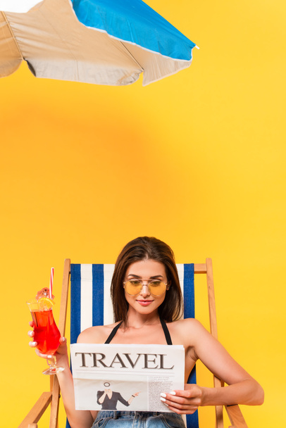 belle femme assise sur une chaise longue avec cocktail tout en lisant le journal de voyage près du parasol de plage sur jaune - Photo, image