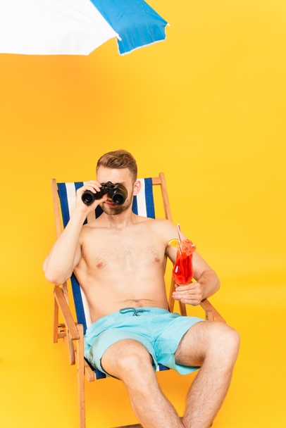 shirtless man holding cocktail and looking through binoculars while sitting on deck chair on yellow  - Photo, Image