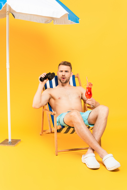 surprised shirtless man holding cocktail and binoculars while sitting on deck chair on yellow  - 写真・画像