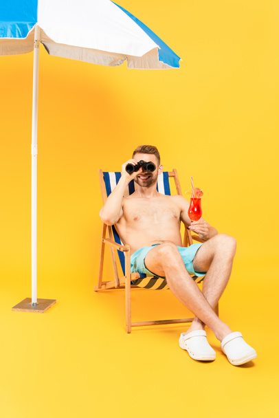cheerful shirtless man holding cocktail and looking through binoculars while sitting on deck chair on yellow  - Фото, зображення