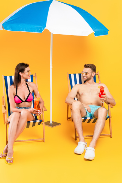 happy couple in sunglasses holding cocktails while sitting on deck chairs near beach umbrella on yellow - Photo, Image