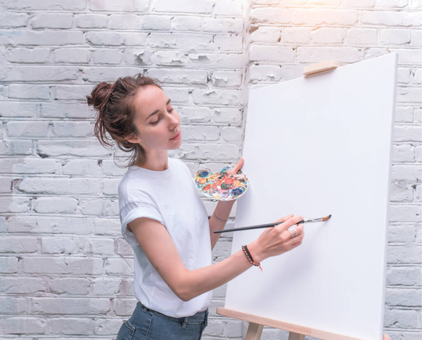 beautiful girl paints picture, artist holds color palette in hand, brush. White blank canvas, easel. Summer in city, brick wall background. Emotions of comfort, pleasure, inspiration and creativity. - Foto, afbeelding