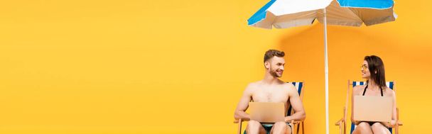 panoramic shot of couple with laptops sitting on deck chairs near beach umbrella on yellow - Photo, Image