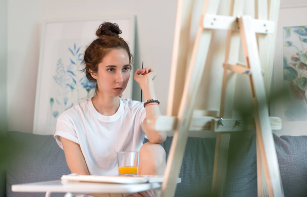 beautiful woman sitting sofa, holding paintbrush, serious look evaluating picture, artist white T-shirt. Easel table. At home in workshop. Emotions fantasy assessment level creativity. Draws picture. - Фото, изображение