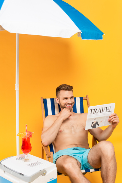 człowiek bez koszulki uśmiecha się czytając gazetę podróżną i siedząc na leżaku obok koktajlu na przenośnej lodówce z zamrażarką na żółto - Zdjęcie, obraz