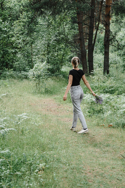young woman with a tail of hair in a black t-shirt and gray jeans in the woods holding blue wildflowers in her hands - Foto, Imagen