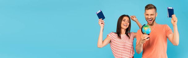 imagen horizontal de pareja feliz sosteniendo pasaportes con boletos aéreos y mirando al globo aislado en azul - Foto, Imagen