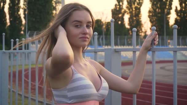 Nahaufnahme Porträt einer jungen schlanken Frau, die nach dem abendlichen Sport in der Nähe des Stadions steht, in die Kamera lächelt und andere zu einem gesunden Lebensstil inspiriert - Filmmaterial, Video