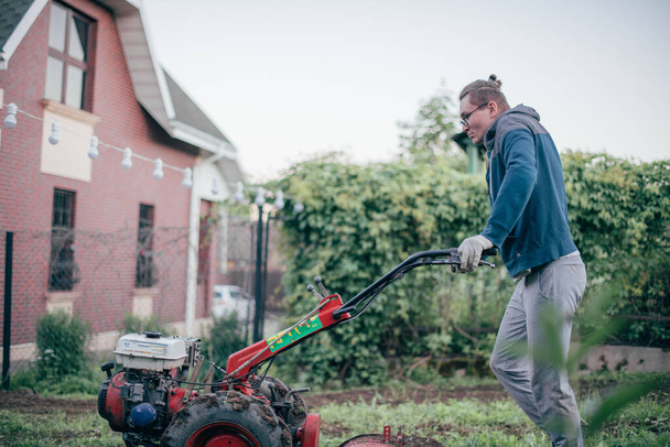 Člověk pracuje na zahradě, obdělává půdu kultivátorem. Mladík s ručním traktorem vykopává půdu pro zeleninovou zahradu ve venkovském domě - Fotografie, Obrázek