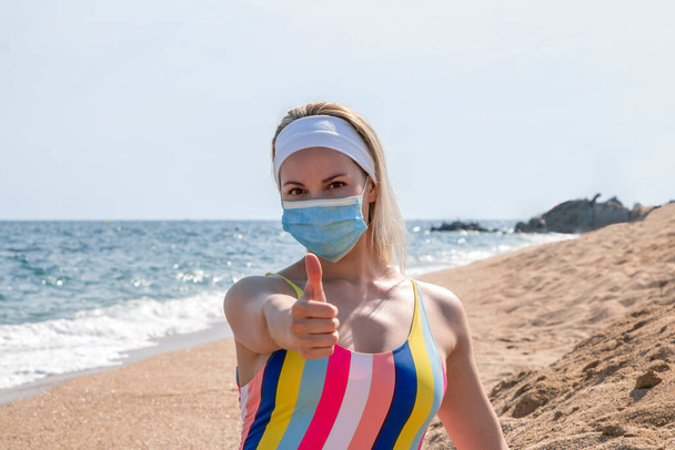 όμορφο κορίτσι με ιατρική μάσκα στην παραλία δείχνει τους αντίχειρες επάνω - Φωτογραφία, εικόνα