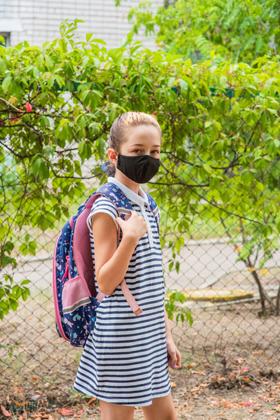 Schoolmeisje meisje met een zwart beschermend masker met een rugzak. Schoolmeisje in schooluniform. Vallen. Binnenkort terug naar school.. - Foto, afbeelding