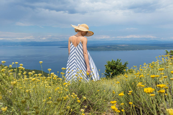 Vista trasera de la mujer joven con vestido de verano a rayas y sombrero de paja de pie en super floración de flores silvestres, relajante mientras disfruta de una hermosa vista de la naturaleza del mar Adriático, Croacia - Foto, Imagen