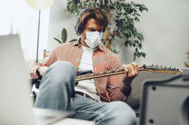 Young man plays guitar at home in medical mask. Coronavirus quarantine lifestyle concept - Foto, Imagem