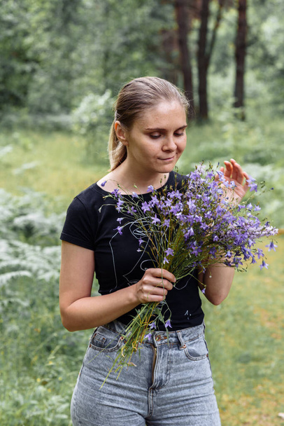 young woman with a tail of hair in a black t-shirt and gray jeans in the woods holding blue wildflowers in her hands - Foto, Imagen