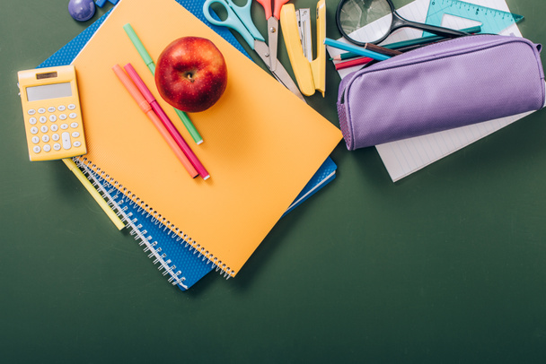 top view of ripe apple and felt pens on multicolored copy books near school stationery on green chalkboard - 写真・画像