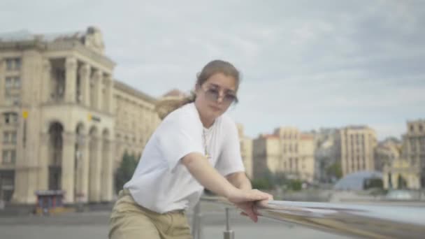Pretty young woman approaching to camera holding handrails and performing robot dance moves. Portrait of cute Caucasian girl in sunglasses dancing with European ancient buildings at the background. - Кадри, відео
