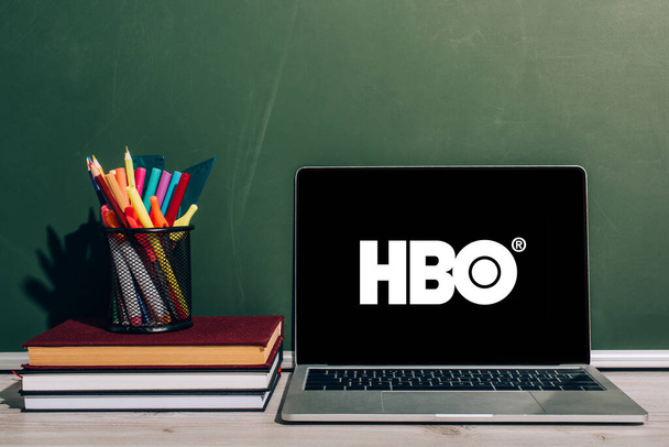 KYIV, UKRAINE - JULY 7, 2020: Laptop with HBO website near pen holder with stationery on stack of textbooks near green chalkboard - 写真・画像