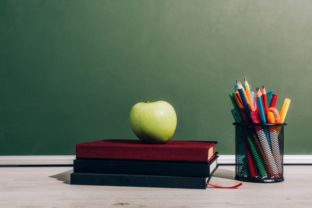 manzana madura en libros cerca del portalápices con útiles escolares en el escritorio cerca de pizarra verde
 - Foto, Imagen