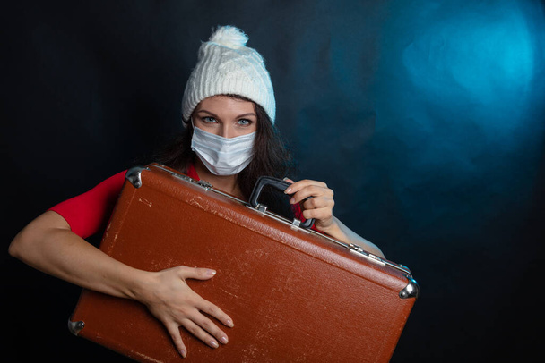 Mladá žena v zimním klobouku a s lékařskou ochrannou maskou drží v rukou na černém pozadí starý kufr. Karanténní koncepce, koronavirus, zimní čas odpočinku - Fotografie, Obrázek
