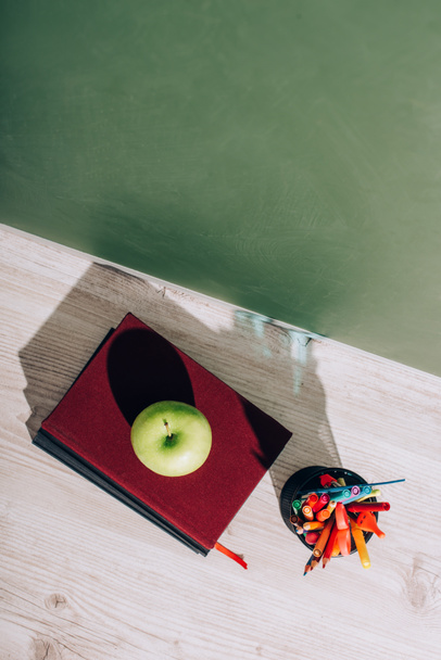 high angle view of whole apple on books near pen holder with stationery on desk near green chalkboard - Photo, Image