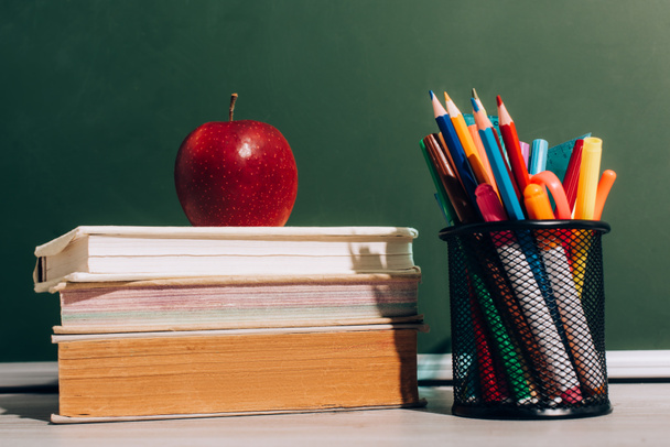 ripe apple on books and pen holder with color pencils and felt pens on desk near green chalkboard - Fotoğraf, Görsel