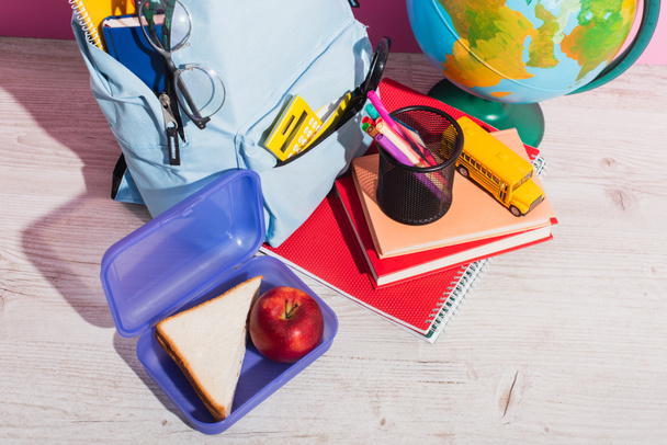 vista de ángulo alto de la mochila azul con útiles escolares cerca de la lonchera, globo, libros, modelo de autobús escolar y manzana en rosa
 - Foto, Imagen
