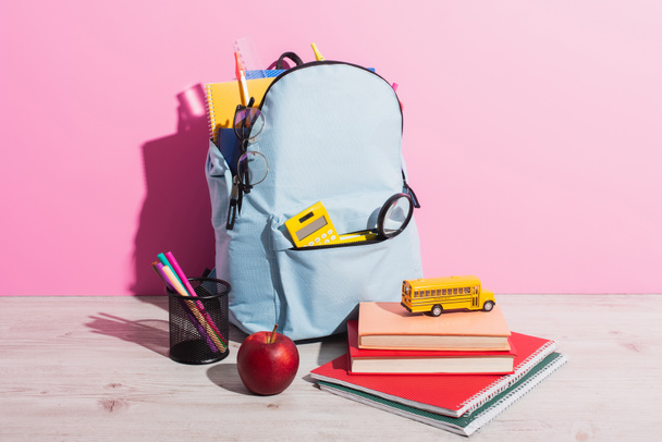pełny plecak z papeterią szkolną w pobliżu autobusu szkolnego zabawki na książki, dojrzałe jabłko i uchwyt na długopis na różowy - Zdjęcie, obraz
