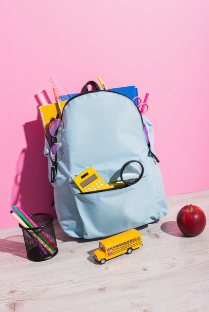 school backpack packed with stationery near school bus model, whole apple and holder with felt pens on pink - Photo, Image